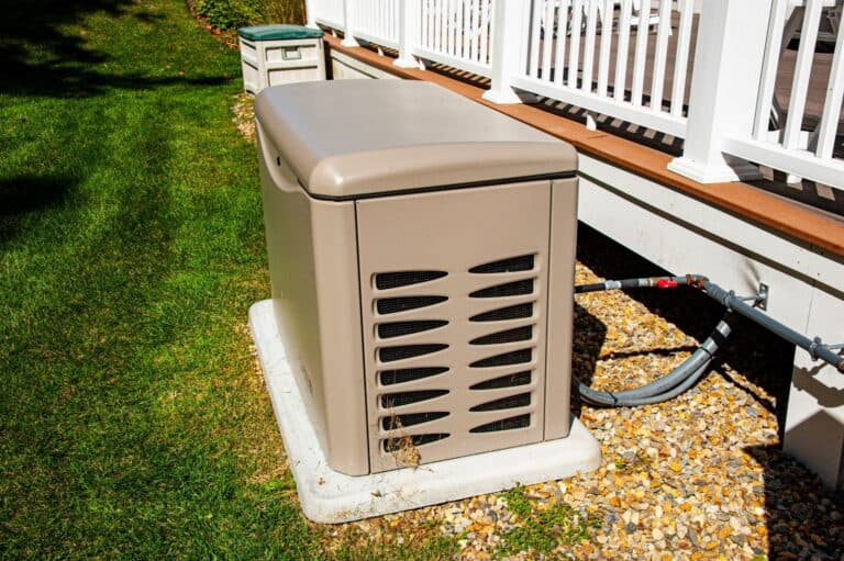 A beige standby generator installation is completed on a concrete pad next to a white railing, with cables connected and surrounded by grass and stones.